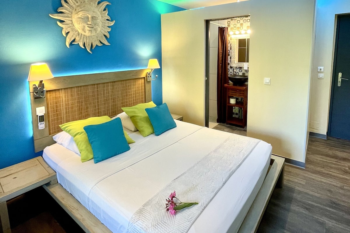 A hotel room with a bed, a nightstand, a wall-mounted lamp, a sun decoration, and a bathroom visible through an open door.