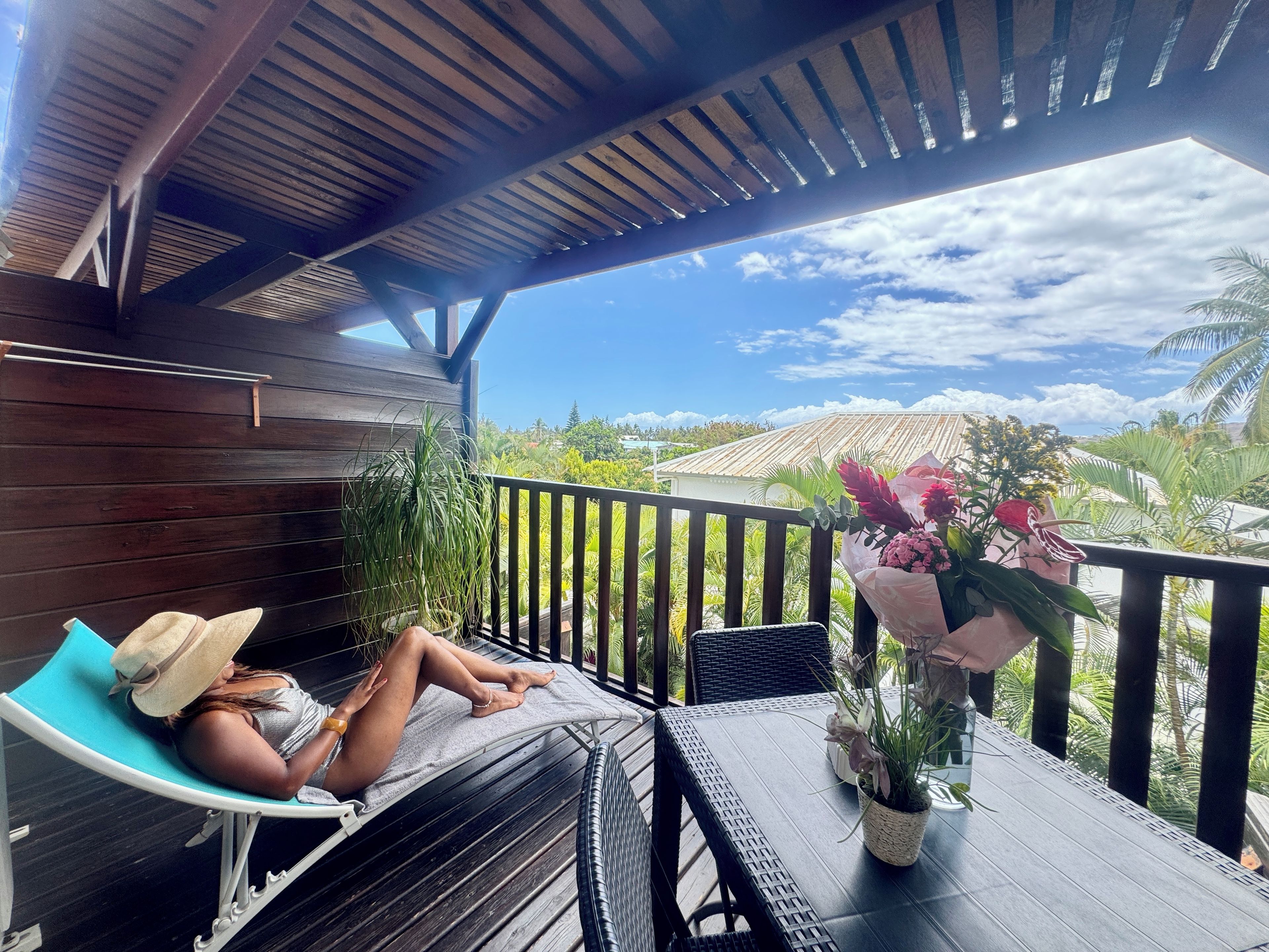Een overdekt terras met een persoon die ontspant in een loungestoel, een tafel met een plant in pot en uitzicht op het omliggende landschap.