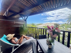 A covered outdoor seating area with a person relaxing on a lounge chair, a table with a potted plant, and a view of the surrounding landscape.
