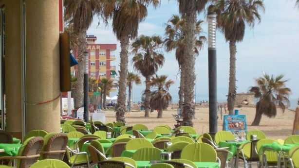 Un área de estar al aire libre con mesas y sillas verdes, palmeras y un edificio con la inscripción 'MAR' al fondo.