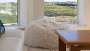 Een kamer met een witte bank, een zachte witte zitkruk en een houten tafel. Een groot raam geeft uitzicht op een groen landschap.