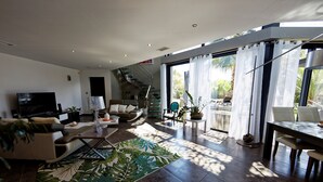 A modern living room with a flat-screen TV, comfortable seating, a staircase, and large windows.