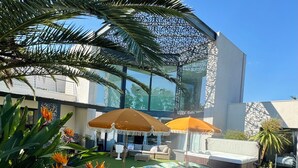 A modern outdoor pool area with lounge chairs and umbrellas, surrounded by tropical plants and a contemporary building.