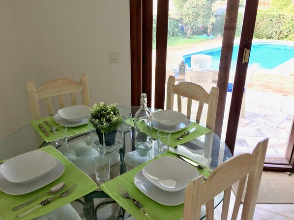 A dining table set for four with green placemats, white plates, and silverware. A small potted plant is on the table, and a glass bottle is visible. The table is near a sliding glass door with a view of a swimming pool. - Sunny villa with pool, 3 bed 3 bath, sea views (Marbella, Málaga, Spain)