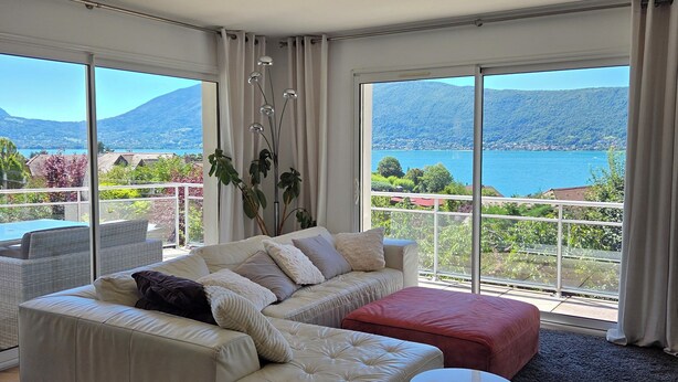 Un salón espacioso con un sofá grande, un reposapiés rojo y una mesa de centro redonda. La habitación tiene ventanas amplias que ofrecen vistas a un lago y montañas.