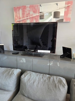 Ein modernes Wohnzimmer mit einem großen Flachbildfernseher an einem grauen Schrank, einer Couch und einem kleinen Tisch mit einer Fernbedienung.