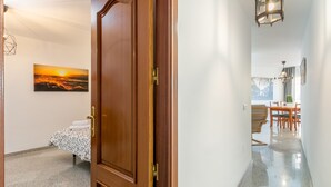 A hallway with a wooden door, a bed with a patterned bedspread, a painting of a sunset, and a dining area with chairs and a table. - SF CARIHUELA LUXURY SUITE (Torremolinos)