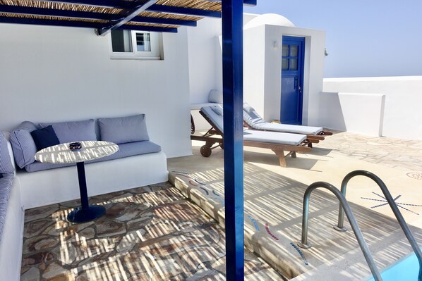 A sunlit outdoor seating area with a stone floor, a blue door, and a pool with metal railings.