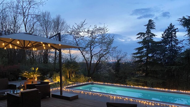 Un área junto a la piscina con una terraza de madera, sillas de descanso e iluminación con luces colgantes.