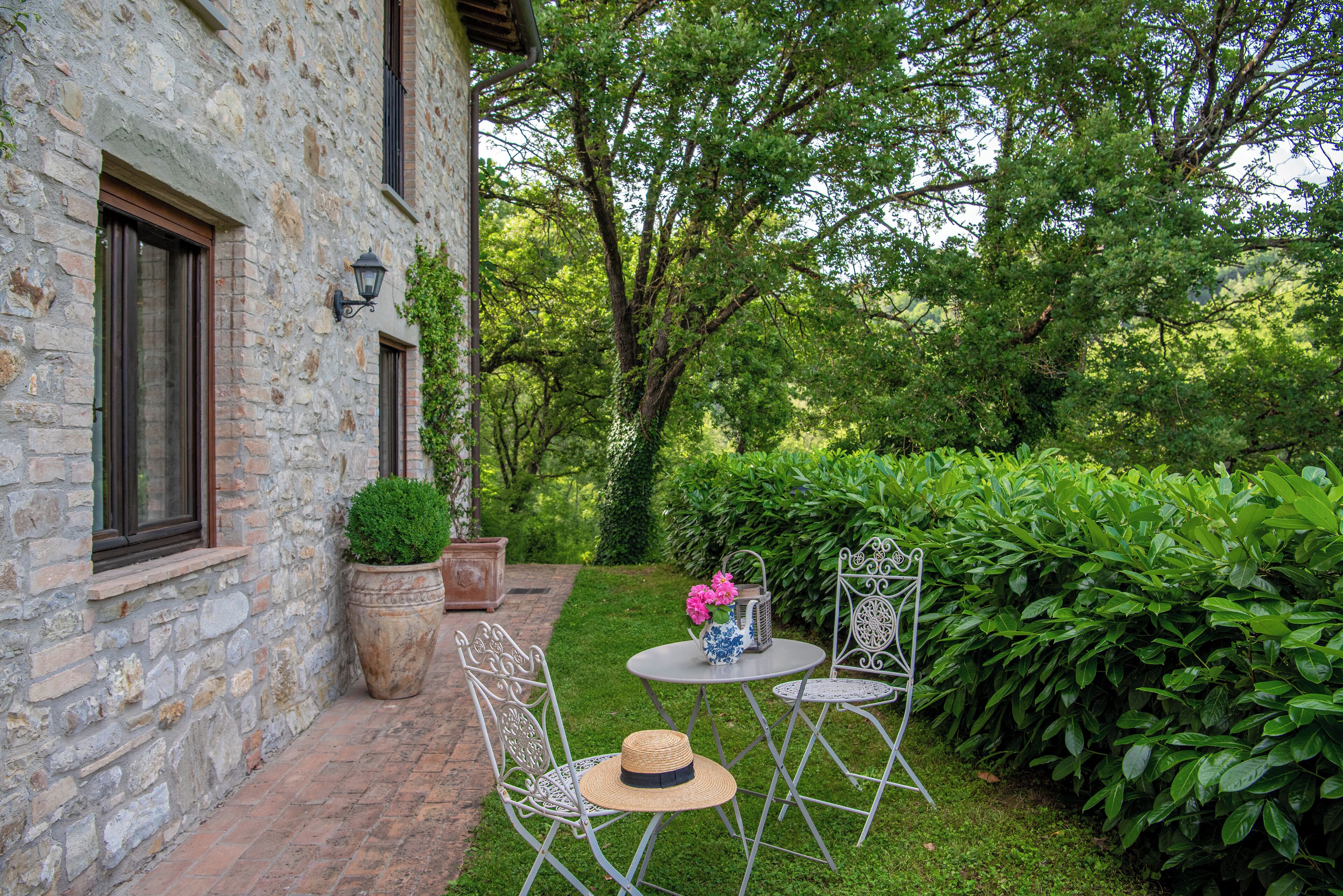 A stone building with a brick pathway leading to a small garden area with metal chairs, a table, and potted plants.