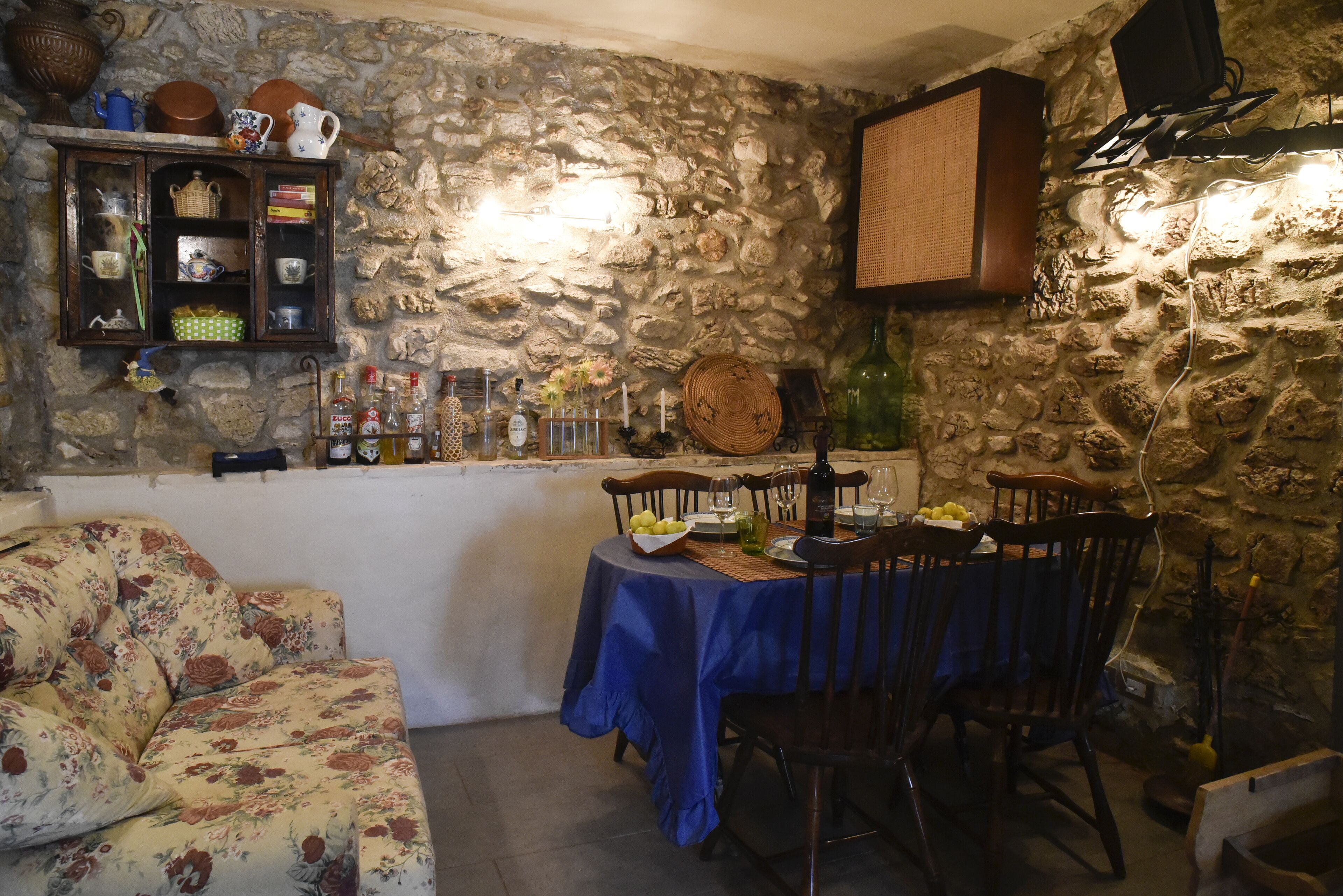 A stone-walled room with a dining table set for two, a floral sofa, and a shelf with various items.