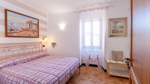 A bedroom with a bed, a nightstand, a chair, a window with lace curtains, and two wall-mounted lamps. - Romantic Seaview Double Room (Corniglia)