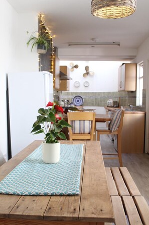 Une table à manger en bois, agrémentée d’un set de table à motifs et d’une plante en pot. On aperçoit en arrière-plan une cuisine avec un réfrigérateur blanc, des placards en bois et un luminaire suspendu.