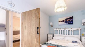 Et soveværelse med dør i træ, seng med hvide sengetæpper, et natbord, en vægmonteret lampe og et maleri af et bybillede.