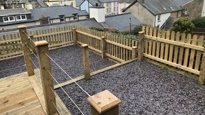 En træterrasse med en grussti, omgivet af et træhegn, med udsigt over et boligområde med huse og en bakke i baggrunden.