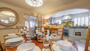 A dining area with a wooden table set for a meal, a chandelier, and a view into the kitchen. - Whitehaven, Brancaster, Norfolk (King's Lynn)