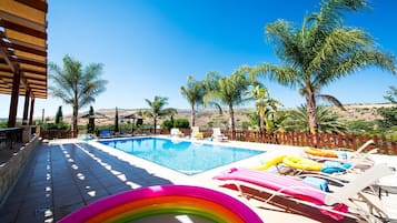 Relax by the serene pool surrounded by palm trees and distant hills, with lounge chairs and loungers for comfortable sunbathing.