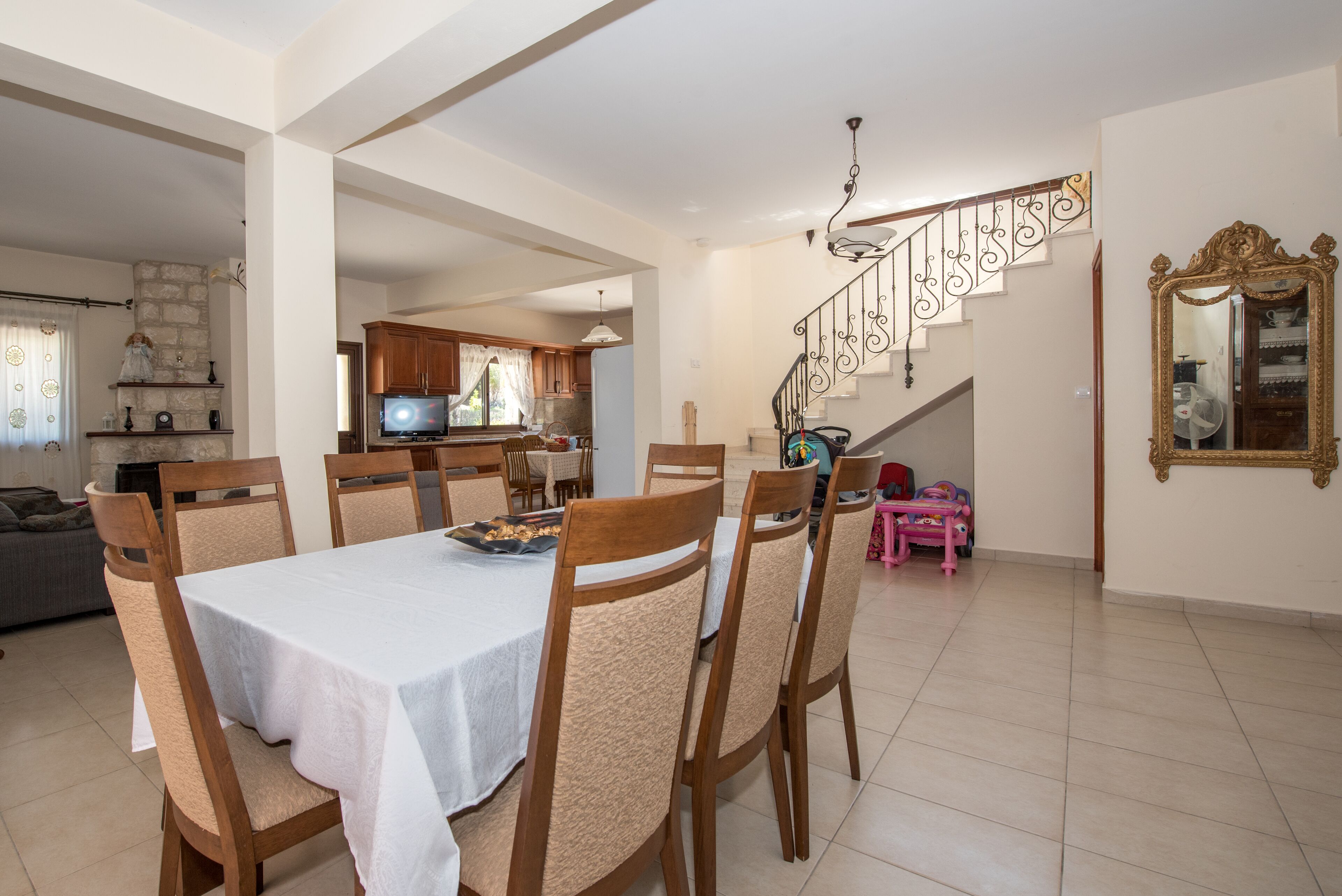 Un coin repas avec une table dressée pour six personnes, des chaises en bois, un téléviseur en arrière-plan, un escalier agrémenté d’une rampe décorative et un grand miroir au mur.