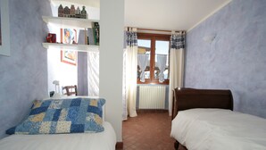 Een slaapkamer met twee bedden, een raam met gordijnen, een radiator, en planken met boeken en decoratieve spullen.
