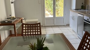 A modern kitchen with white cabinets, a stainless steel sink, and a built-in oven. There is a dining area with a glass table and chairs, and a small potted plant on the table.
