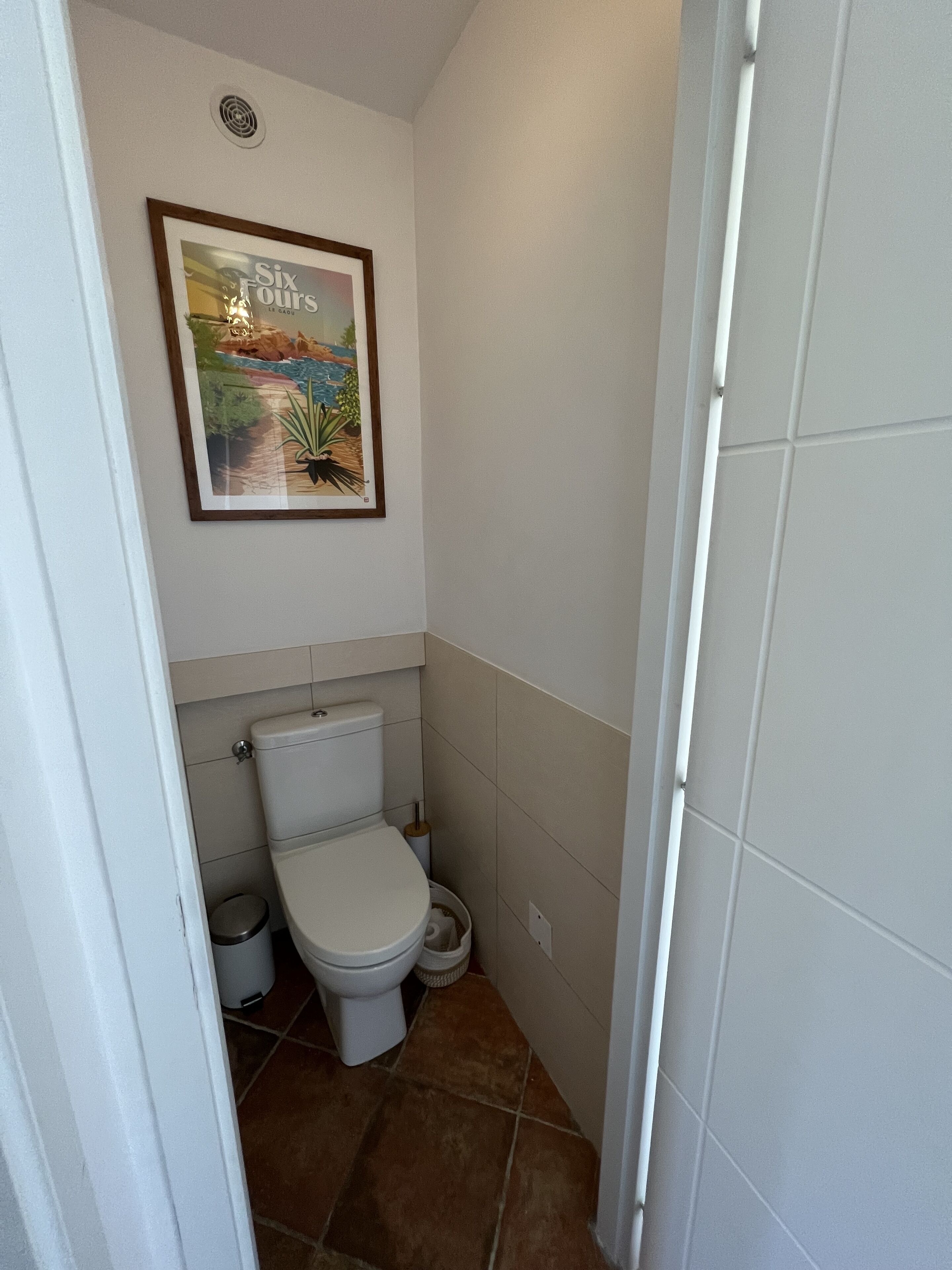 Un baño pequeño y limpio, con inodoro, un cubo de basura y un cuadro en la pared.
