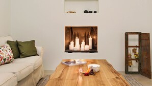 Uma sala de estar com mesa de centro de madeira, um sofá com almofadas verdes, lareira com velas e uma pequena estante com objetos decorativos.