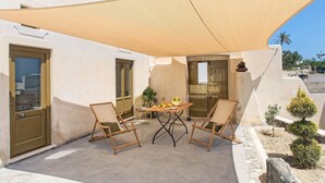 Um pátio externo com uma mesa posta para dois, cercado por cadeiras de madeira, uma grande vela de sombreamento e um pequeno jardim com pedras e plantas.