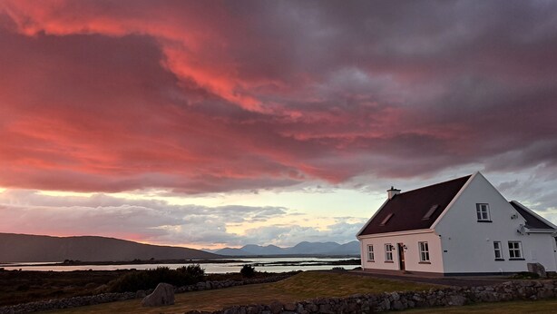 A white house with a sloped roof, situated on a grassy area with a stone wall in the foreground. - Luxury Connemara Cottage With Panoramic Views Of The Sea And Mountains  (Rosmuc)
