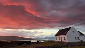 A white house with a sloped roof, situated on a grassy area with a stone wall in the foreground. - Luxury Connemara Cottage With Panoramic Views Of The Sea And Mountains (Rosmuc)