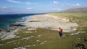 A person standing on a grassy hill overlooking a beach with a clear sky. - Luxury Connemara Cottage With Panoramic Views Of The Sea And Mountains  (Rosmuc)