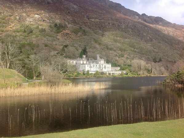 A historic building with a lake in the foreground and a mountainous backdrop. - Luxury Connemara Cottage With Panoramic Views Of The Sea And Mountains (Rosmuc)