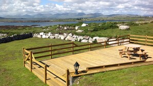 A wooden deck with picnic tables and a lantern, overlooking a lake and mountains. - Luxury Connemara Cottage With Panoramic Views Of The Sea And Mountains  (Rosmuc)