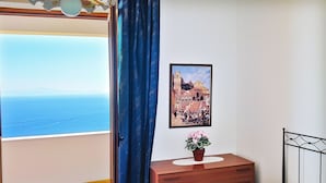 Une chambre à coucher avec un lit, une commode en bois, un tableau encadré au mur et une vue sur la mer par une porte ouverte.