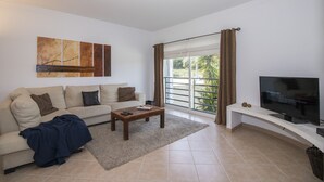 Una sala de estar con sofá, mesa de centro, televisor y un amplio ventanal con cortinas.
