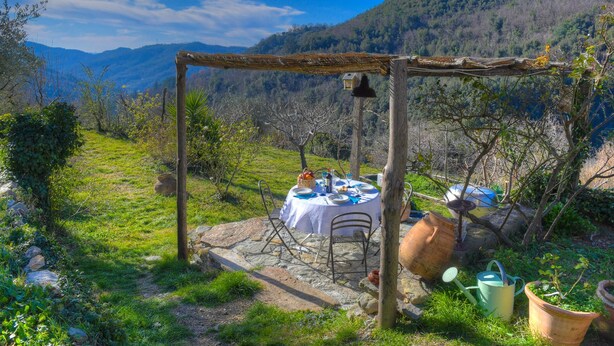 Ein rustikales Essensemble im Freien mit Tisch und Stühlen unter einer Holzpergola, umgeben von Grünpflanzen und einer Bergkulisse.