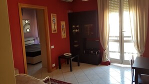 Ein Zimmer mit Bett, Fernseher, einem kleinen Tisch und einer Couch. Es gibt ein Fenster mit Vorhängen, eine Klimaanlage und eine Tür zu einem anderen Raum.