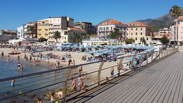 Ein Strand mit einer Holzpromenade, Sonnenschirmen und Menschen, die das Meer genießen.