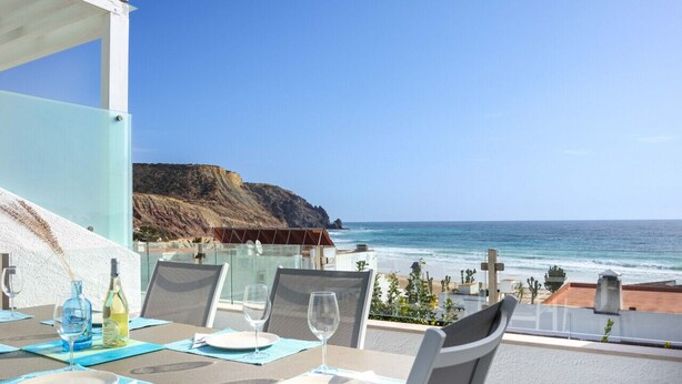 A dining table set with plates, glasses, and a bottle, overlooking a beach with cliffs and the ocean. - Beco do Poço - Praia da Luz (Luz)