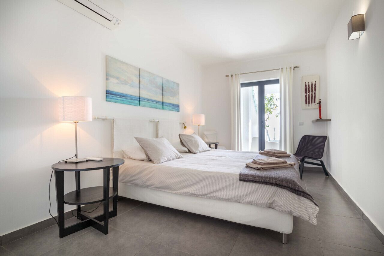 A modern bedroom with a large bed, two bedside tables with lamps, a chair, and artwork on the walls.