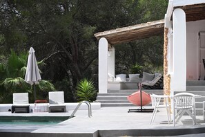 Uma área de piscina com cadeiras de lounge brancas, um guarda-sol vermelho e uma área de estar coberta com almofadas.