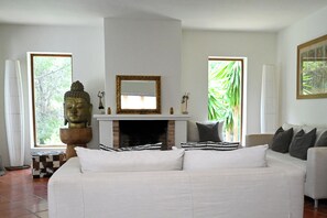 Sala de estar com sofá branco, estátua de Buda, lareira e janelas amplas com vista para o verde.