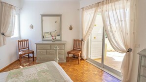 Ein Schlafzimmer mit einem Bett, zwei Holzstühlen, einem Schminktisch, einem Spiegel und einer Glastür zum Außenbereich.