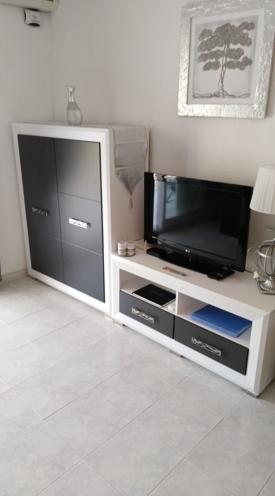 A modern living room with a flat-screen TV on a white entertainment unit, a cabinet with drawers, and a framed picture on the wall.