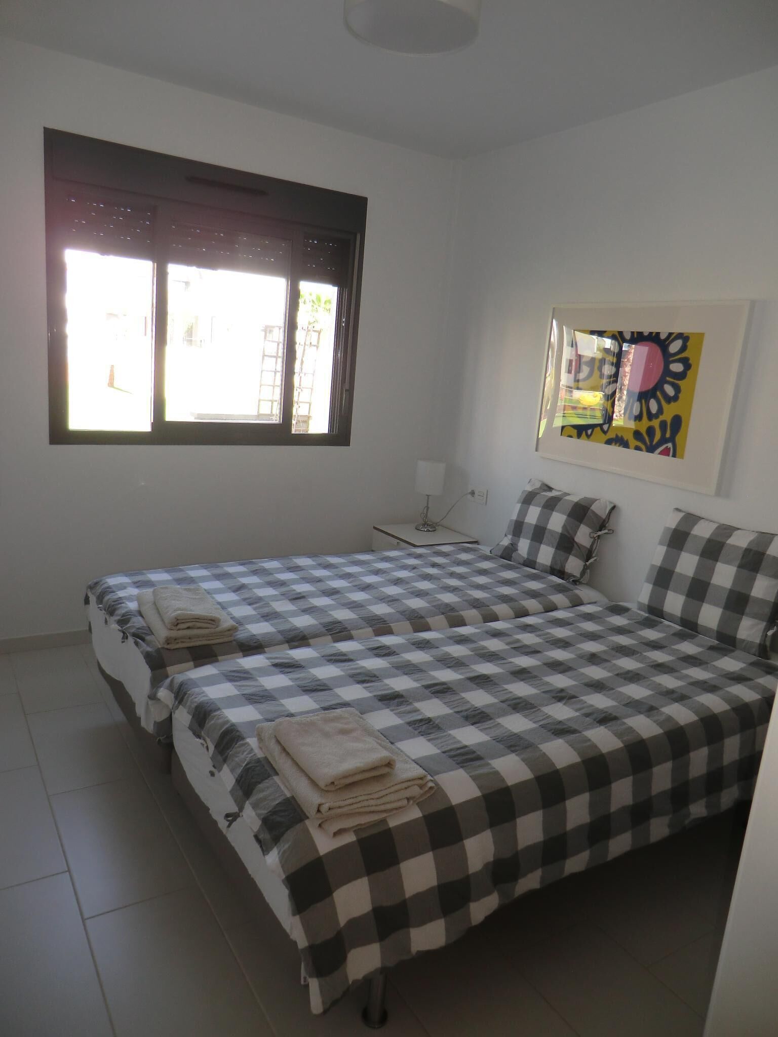 A room with two beds, a window, a bedside table, and a wall-mounted artwork.