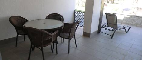 A covered patio area with a round table and four wicker chairs, a metal chair, and a view of palm trees and buildings in the background.