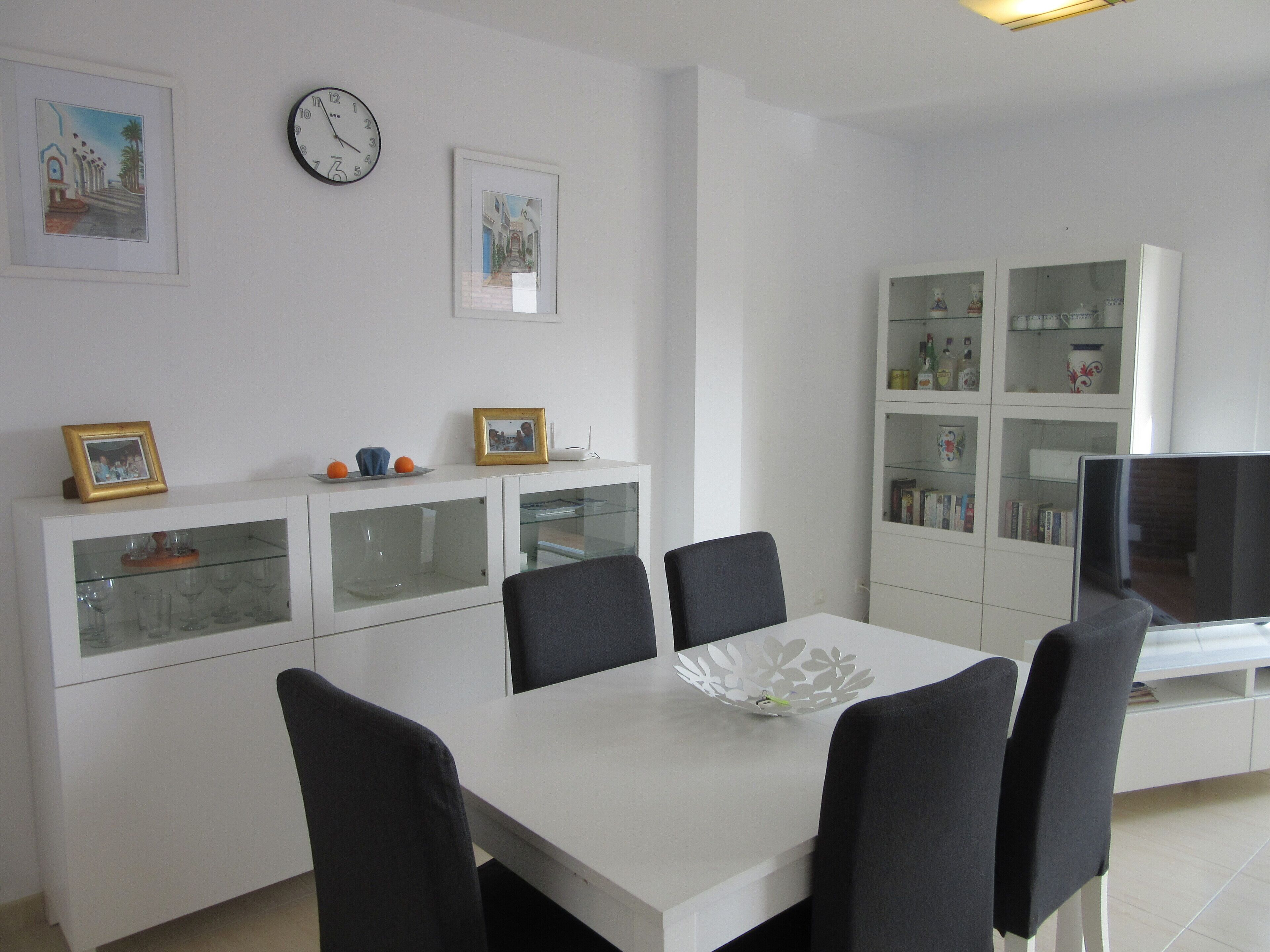 Una sala moderna con una mesa de comedor blanca, sillas negras, un aparador de cristal con objetos decorativos y un televisor sobre un mueble blanco.