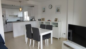 A modern kitchen with white cabinets, a dining area with a white table and black chairs, a large flat-screen TV, and a clock on the wall.