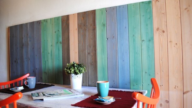 Un coin repas avec un mur en bois peint de différentes couleurs, une table avec un set de table rouge, deux chaises orange, une tasse bleue, une tasse blanche, une petite plante en pot et un magazine.