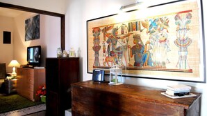 Une pièce comprenant un grand tableau sur le thème de l’Égypte ancienne, une commode en bois, un téléviseur et une lampe.