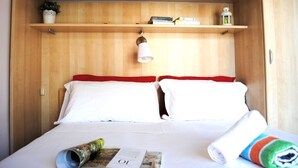 Un lit bien fait, avec du linge de lit blanc et des touches de rouge, une petite étagère avec des livres et une lanterne, et un magazine enroulé posé sur le lit.